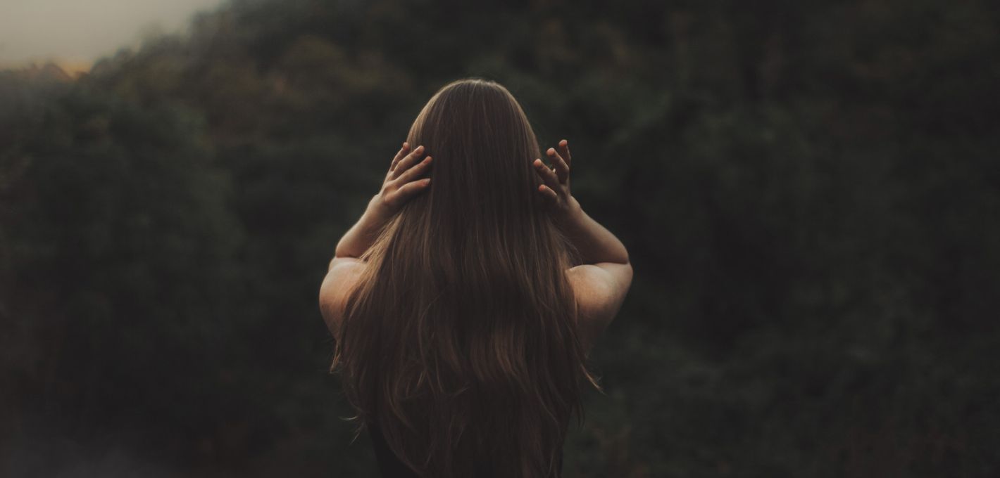 Frau mit langen Haaren von hinten, die Hände am Kopf, vor dunklem Wald als Bild für das Thema Nervensystem regulieren